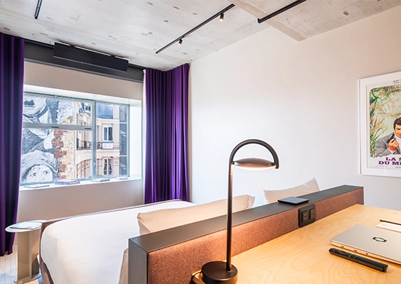 Espace nuit avec lit double, coin bureau intégré en tête de lit et rideaux violets près d'une fenêtre donnant sur la ville.