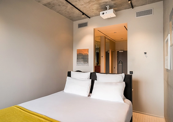 Chambre d'hôtel épurée avec lit double, draps blancs, plaid jaune et projecteur fixé au plafond.