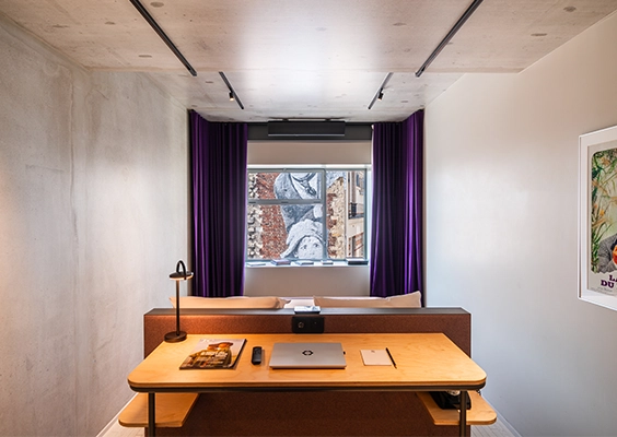 Espace de travail moderne aménagé avec bureau en bois, lampe posée et vue sur le lit encadré de rideaux violets.