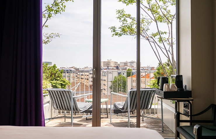 Hotel room with a floor-to-ceiling window opening onto a balcony overlooking the city rooftops, with a kettle tray nearby