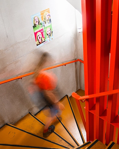 Blurred silhouette rushing down a spiral staircase with yellow steps and a bright red structure, colorful movie poster on the wall