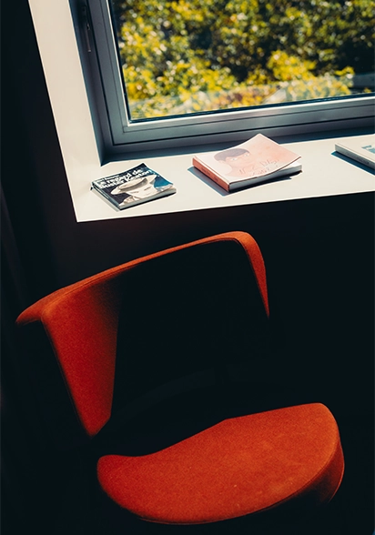 Bright orange designer armchair beneath a window ledge stacked with magazines, overlooking backlit greenery