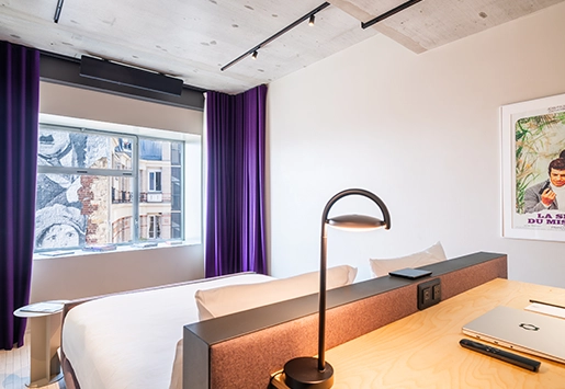 Bedroom with a raw concrete ceiling and purple curtains, a white bed against a terracotta headboard, and a light wood desk