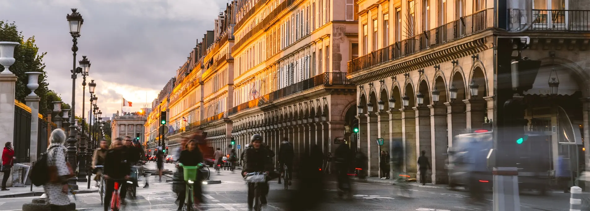 Cyclists and pedestrians streaming along Rue de Rivoli bathed in golden light, with arches and streetlamps in the foreground