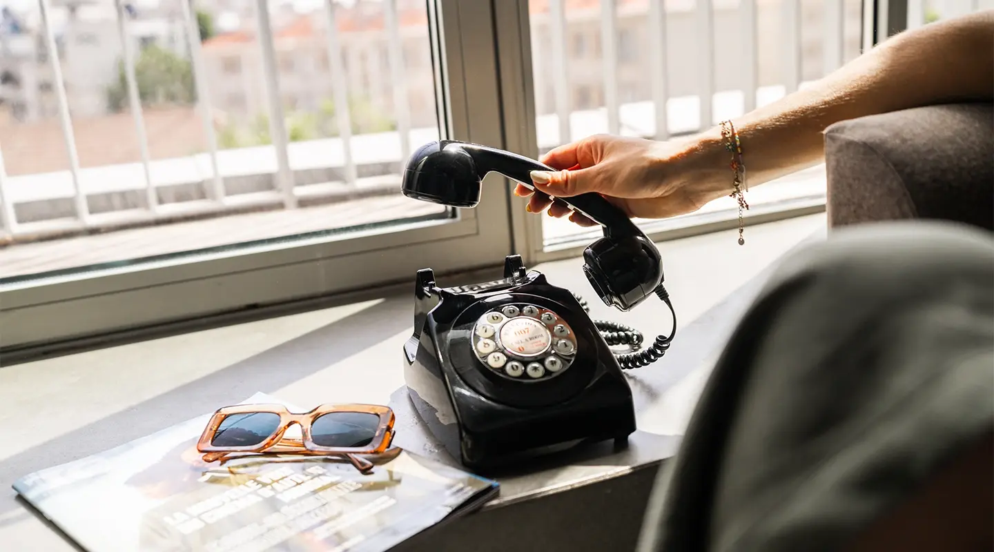 Vue en format portrait d'une femme décrochant un téléphone rétro sur un rebord de fenêtre, avec magazine et lunettes de soleil