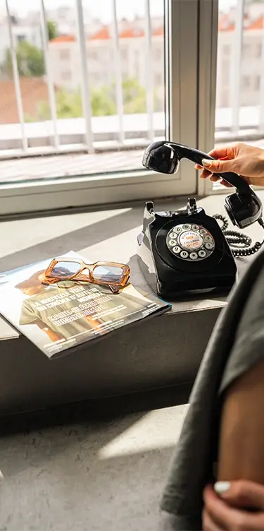 Vue en format portrait d'une femme décrochant un téléphone rétro sur un rebord de fenêtre, avec magazine et lunettes de soleil