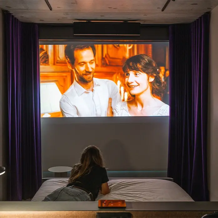 Spectatrice allongée sur un lit double regardant un film projeté sur grand écran entre des rideaux violets