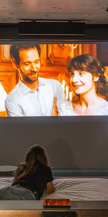 Woman lying on a bed in the foreground watching a movie projected onto a large wall screen