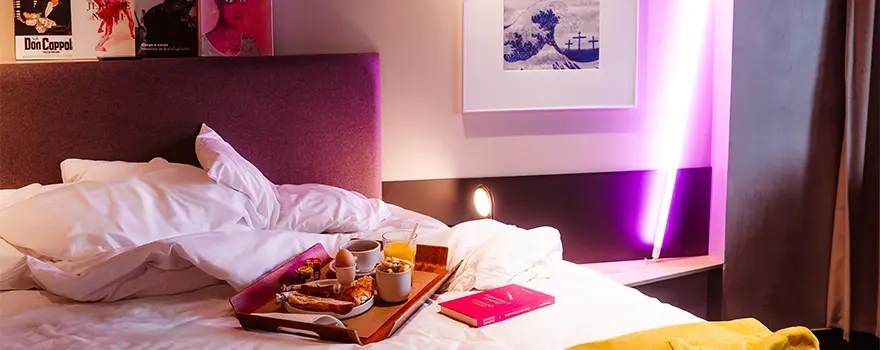 Plateau de petit-déjeuner gourmand posé sur un lit défait avec un livre dans une chambre d'hôtel éclairée au néon rose