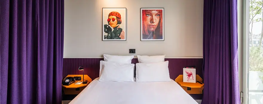 Chambre d'hôtel design avec lit double, rideaux violets et tableaux d'art moderne au-dessus de la tête de lit