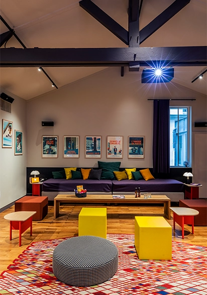 Colorful living room under a wooden frame, featuring a purple bench with green and yellow cushions, cube ottomans, and framed movie posters
