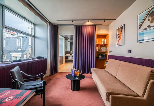 Modern hotel room with a minimalist design, featuring a sofa, a coffee table with snacks, and a large window overlooking the rooftops.