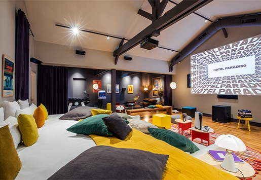 Spacious cinema-style loft with exposed beams, featuring a massive bed piled with colorful cushions facing a large projection screen.