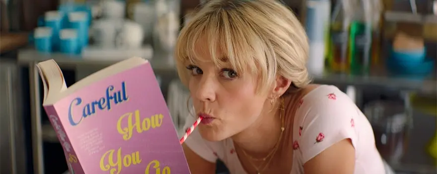 Femme blonde avec une frange qui lit un livre avec une paille dans la bouche