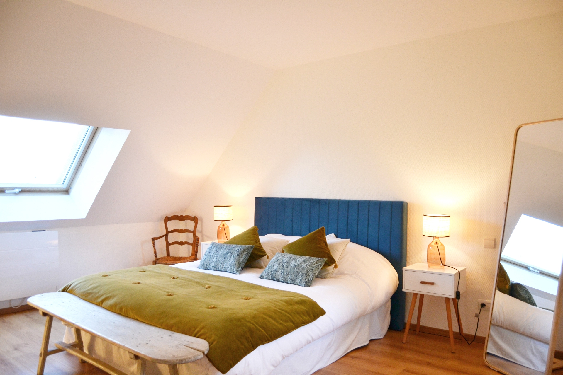 A bright master bedroom in the attic with a double bed, a blue headboard, a Velux skylight and a full-length mirror.