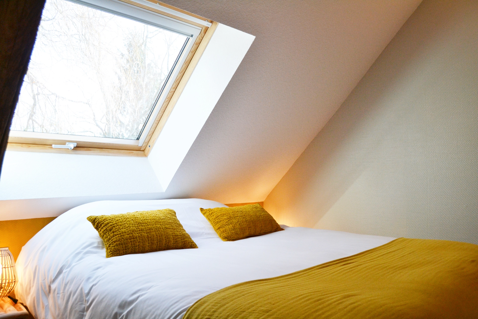A comfortable double bed beneath a Velux skylight, complete with cushions and a mustard-yellow bedspread, in an attic bedroom.