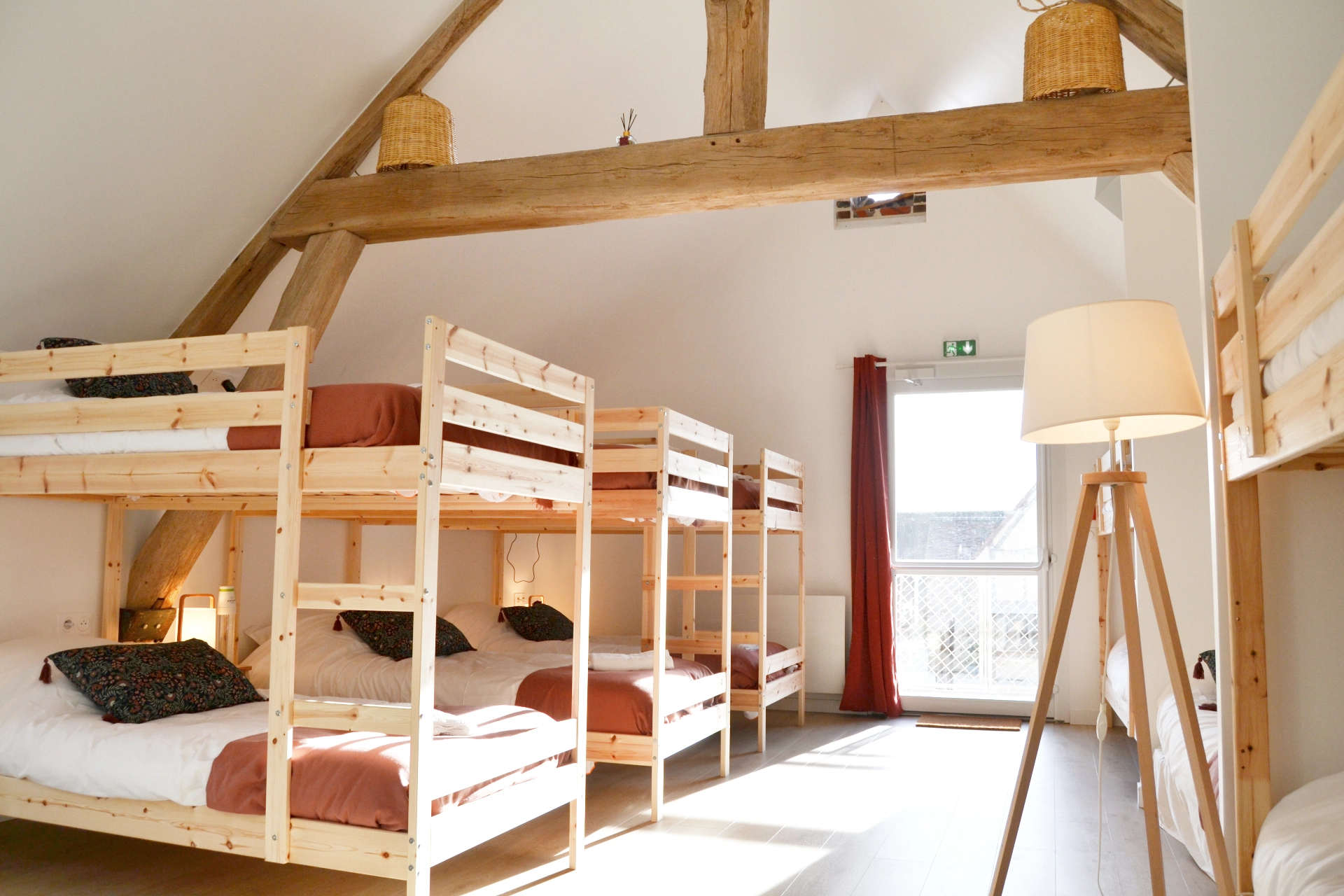 A spacious, bright dormitory with light-coloured wooden bunk beds, exposed timber beams and a stylish tripod floor lamp.