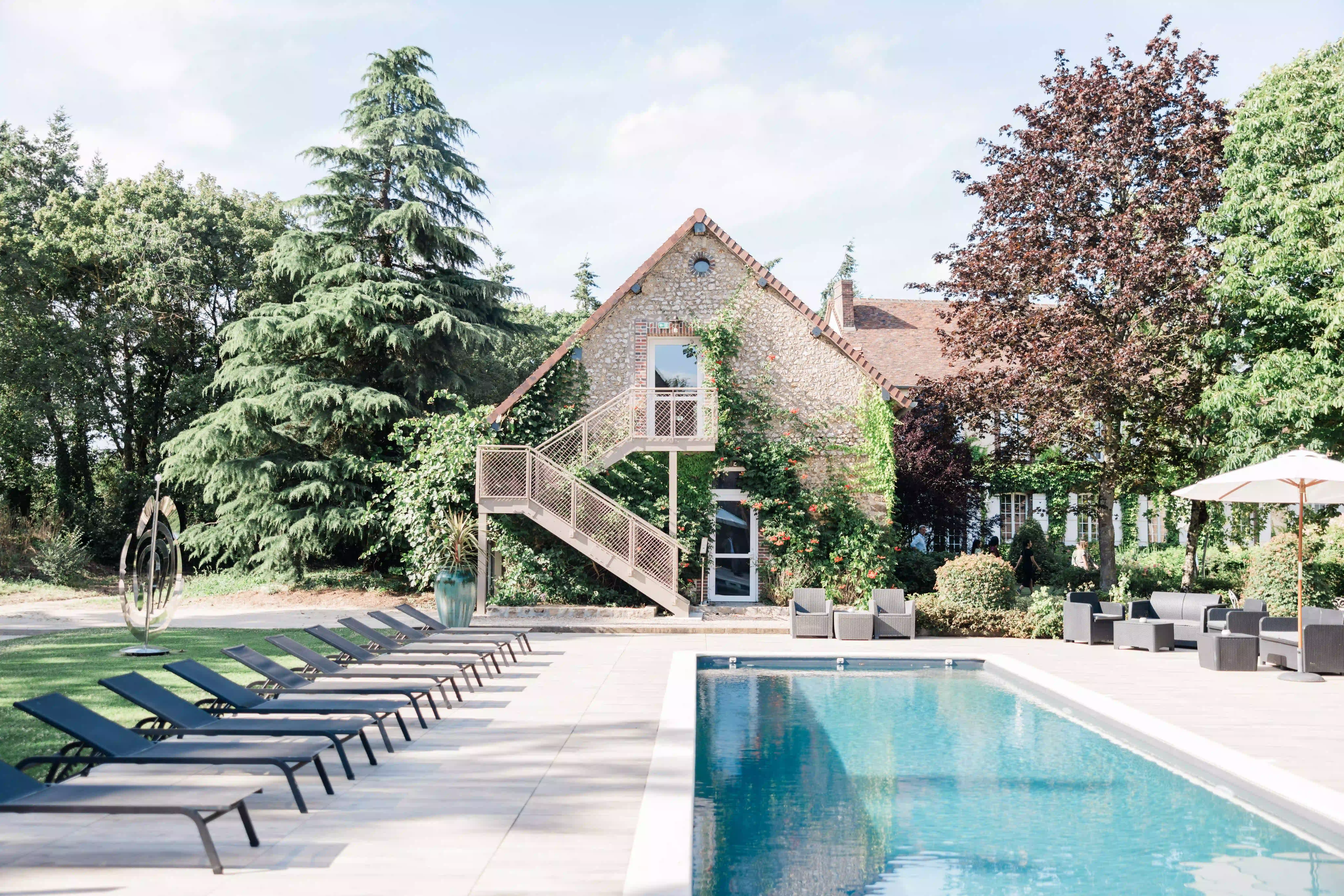 A rectangular outdoor swimming pool with sun loungers and a tree-lined garden in front of a large, charming stone building.