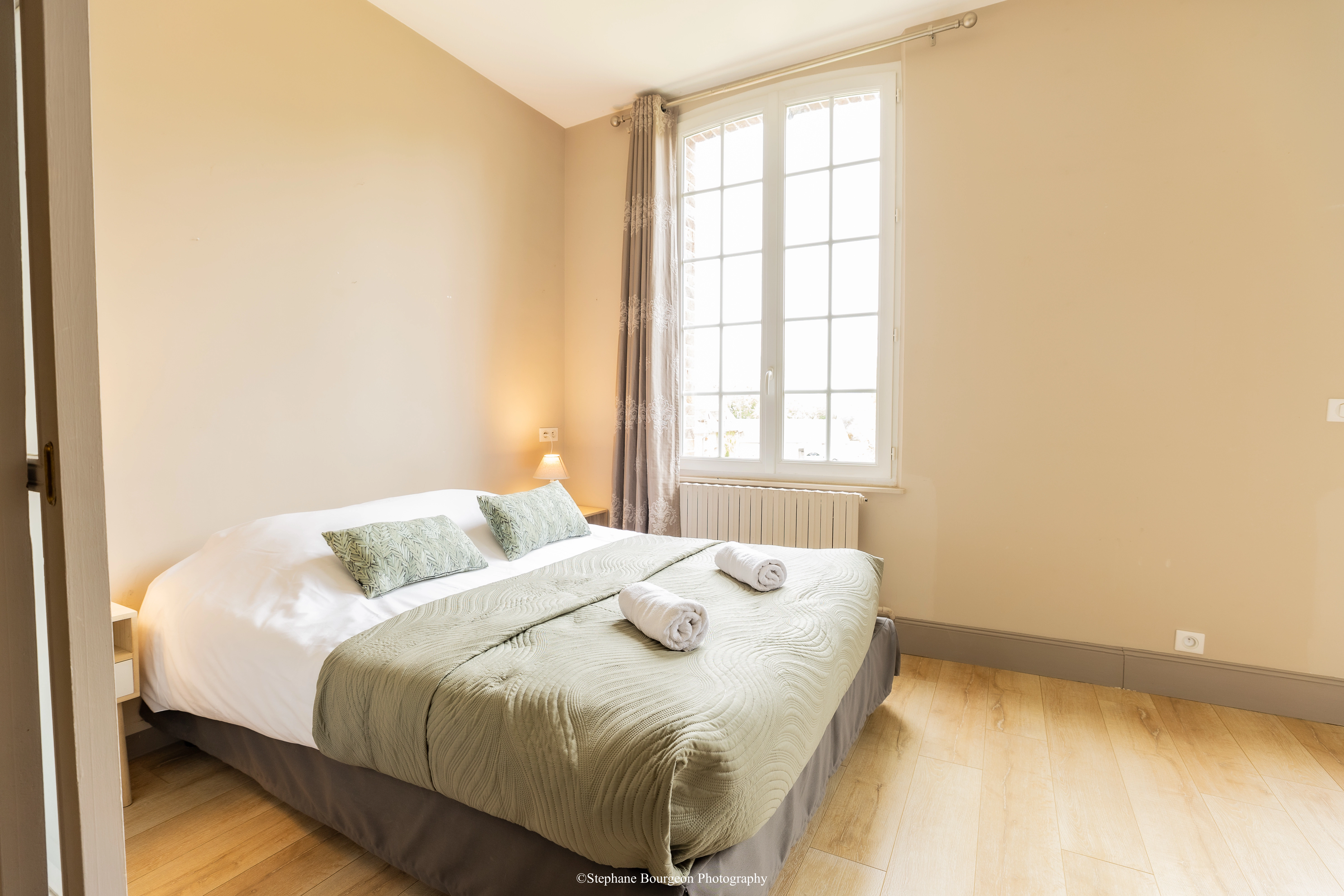 An elegant and bright double room with a made-up bed, light grey bed linen, a large window with small panes, and wooden flooring.
