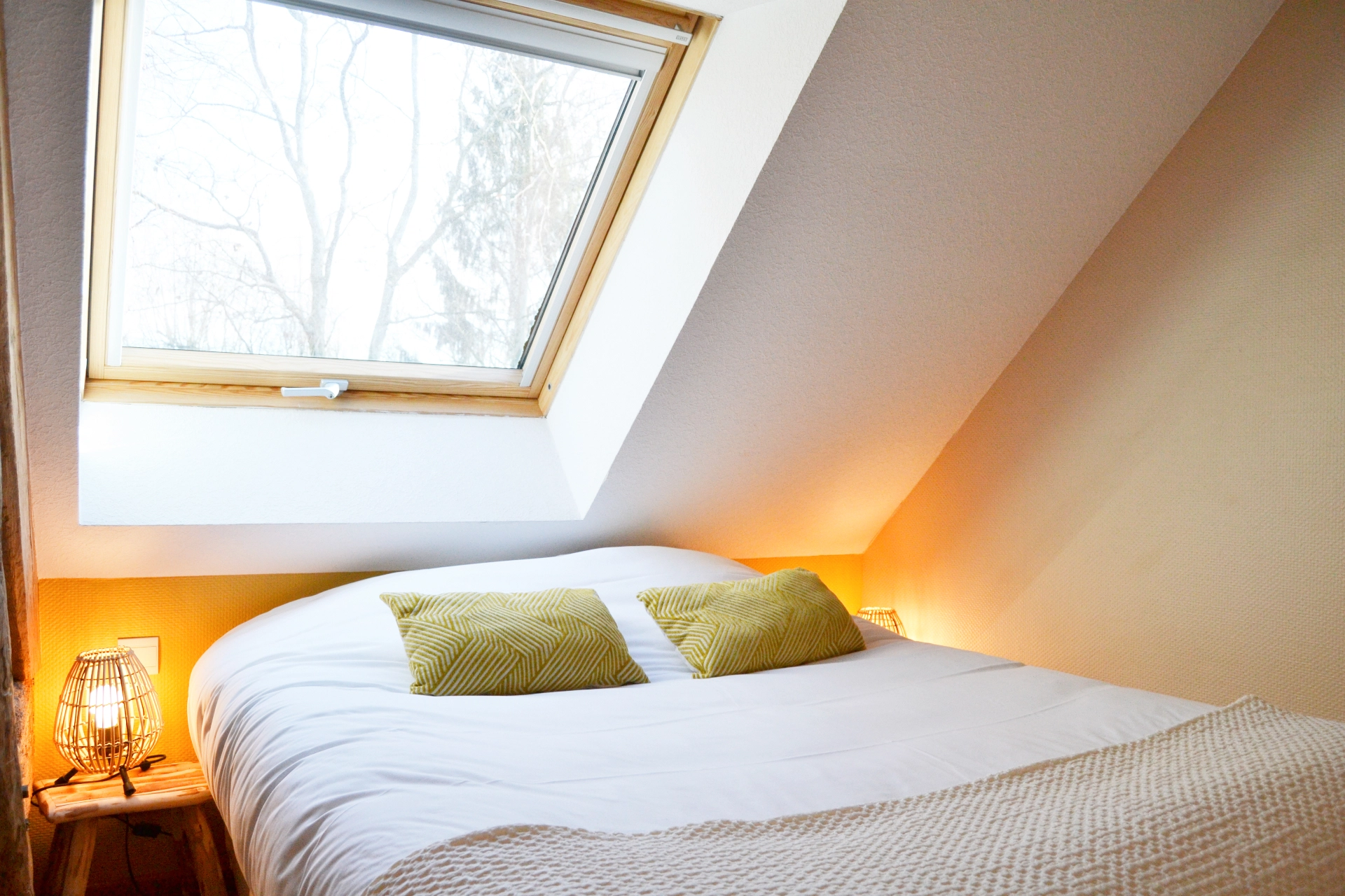 A comfortable double bed beneath a Velux skylight, complete with cushions and a mustard-yellow bedspread, in an attic bedroom.