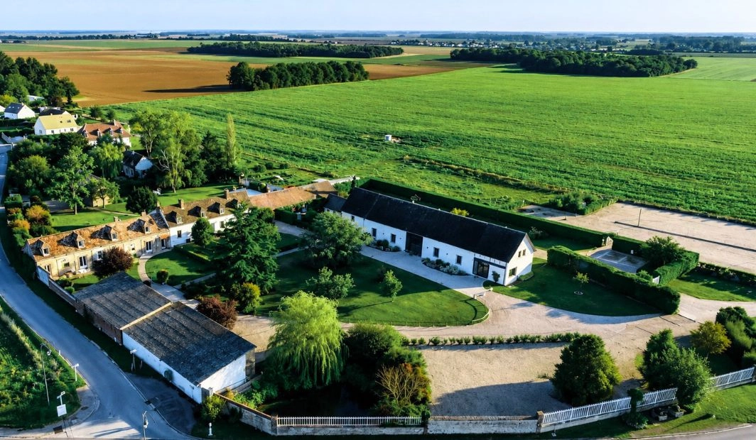 Vue aérienne d'un domaine rural avec grange rénovée blanche, parc arboré et vastes champs agricoles à l'horizon
