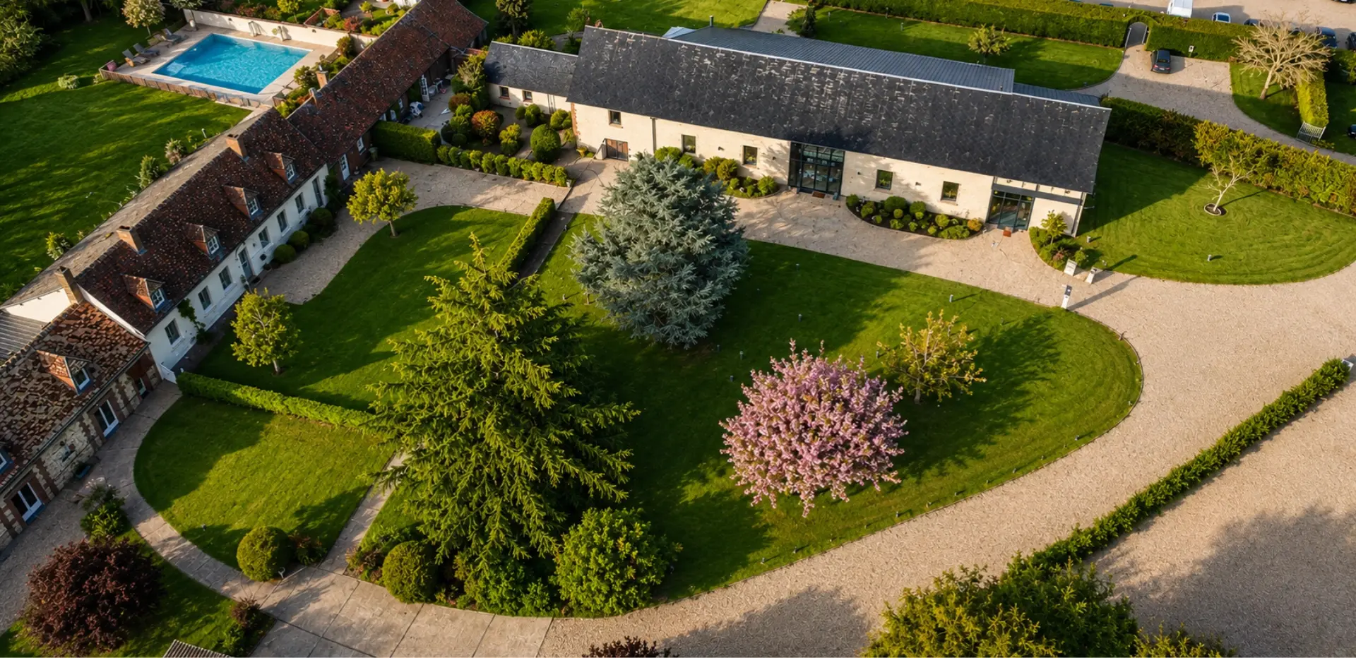 Vue aérienne d'un domaine avec pelouse arborée, bâtiments en pierre, piscine extérieure et allées en gravier