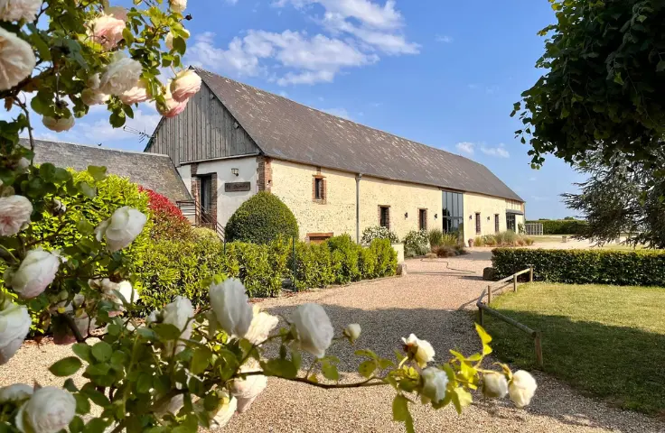 Façade d'une grange en pierre rénovée entourée de jardins soignés, avec rosiers en fleurs au premier plan et ciel bleu