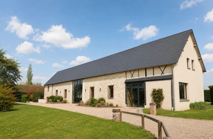 Façade extérieure d'une grange en pierre rénovée avec toit ardoise et grandes baies vitrées, entourée d'une pelouse verdoyante