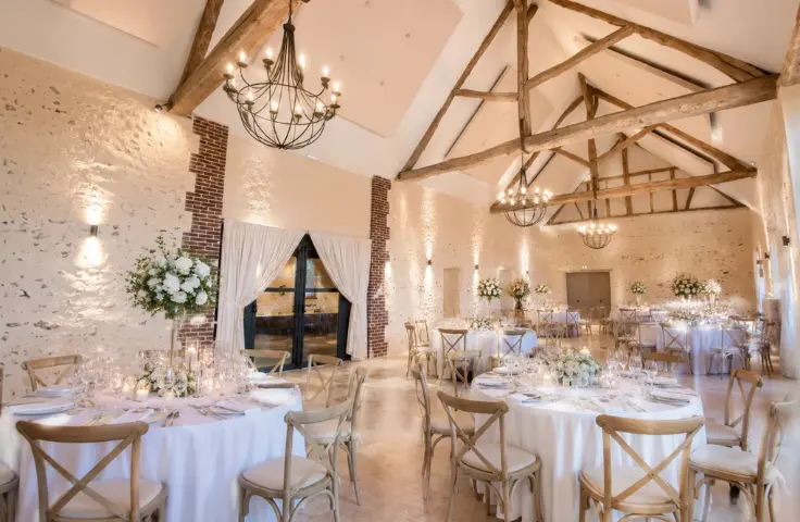 Salle de réception de mariage en grange avec poutres apparentes, lustres en fer forgé, tables blanches dressées et compositions florales blanches