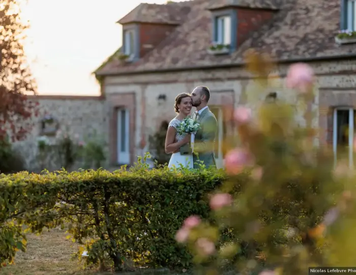 Couple de mariés s'embrassant dans un jardin fleuri devant un bâtiment en pierre et brique au coucher du soleil