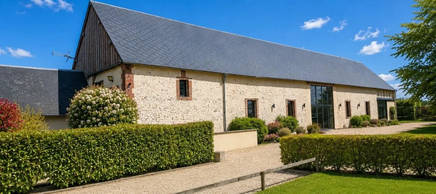 Façade latérale de la grange en pierre avec toit ardoise, grandes baies vitrées, haies taillées et hortensias sous ciel bleu