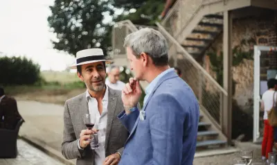 Deux hommes élégants conversant lors d'un événement en plein air, l'un avec un chapeau de paille et un verre de vin