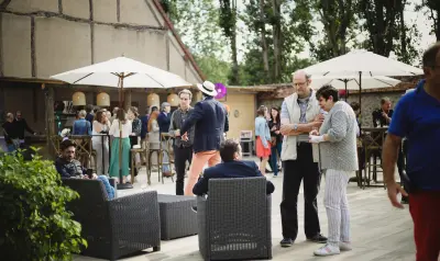 Invités conversant sur une terrasse en bois lors d'un événement en plein air, avec parasols blancs et grange à colombages en arrière-plan