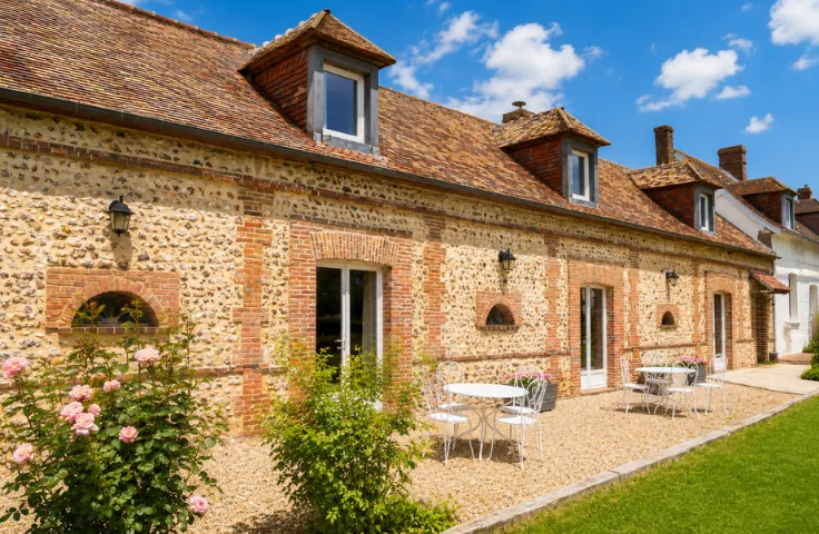 Façade en pierre et briques d'une longère normande avec lucarnes en zinc, terrasse gravillonnée meublée de tables et chaises blanches, rosiers en fleurs.