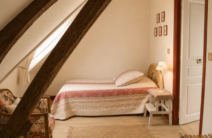Petite chambre mansardée avec lit simple à couverture rayée rose et beige, fauteuil fleuri et poutre en bois traversant la pièce.
