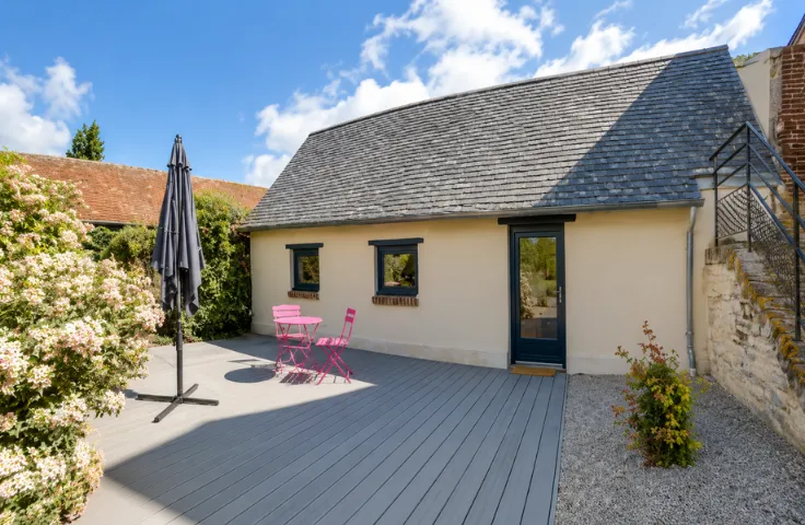 Petite maison avec toit d'ardoise, terrasse en bois avec table et chaises roses, parasol et massif fleuri.