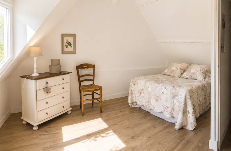 Chambre mansardée lumineuse avec commode blanche ancienne, chaise en bois et paille, lit à couverture fleurie beige et rose.