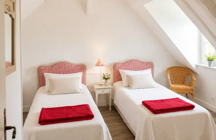 Chambre mansardée avec deux lits jumeaux à têtes de lit à carreaux rouge et blanc, serviettes rouges pliées, fauteuil en rotin près de la fenêtre.
