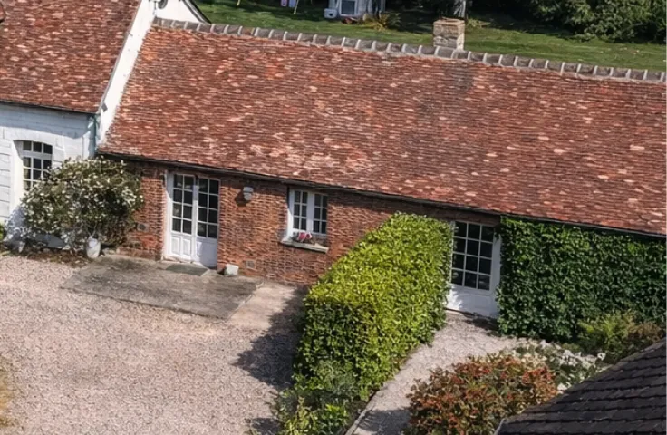 Vue rapprochée d'une toiture en tuiles anciennes et d'une façade en briques avec porte-fenêtres blanches, entourée de haies et d'une allée gravillonnée.