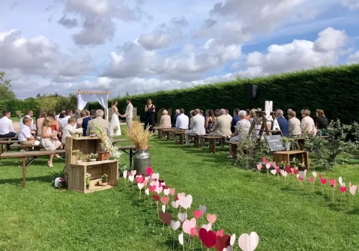 Cérémonie laïque en extérieur sur une pelouse, arche en bois et tissu blanc, allée bordée de cœurs décoratifs roses et rouges, invités assis sur des bancs en bois devant une haie.