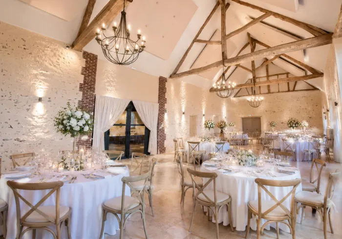 Grande salle de réception en pierre avec lustres en fer forgé, poutres apparentes et tables rondes dressées pour un dîner de mariage.