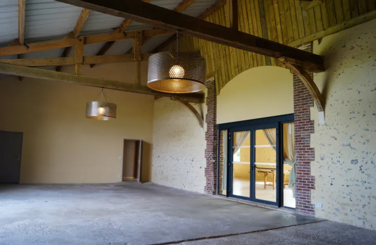 Grand espace intérieur brut à charpente en bois et arche en briques, sol en béton et grande porte-fenêtre ouvrant sur une salle voisine, lustres suspendus en métal ajouré.