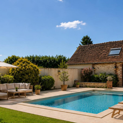 Piscine extérieure entourée de chaises longues, canapé, parasol et murs avec végétation sous un ciel bleu clair.