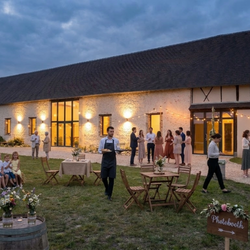 Réception en soirée dans une cour avec invités, tables en bois et décorations florales.