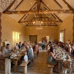 Invités assis à une longue table en bois dans une salle de réception aux murs en pierre, décorée de guirlandes lumineuses.