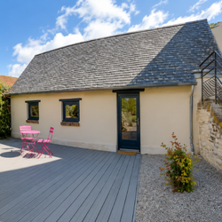 Petite maison beige avec toit en ardoise, porte noire, fenêtres, et mobilier rose sur terrasse en bois.