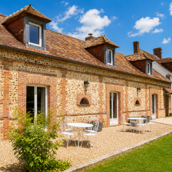 Maison en brique avec toit en tuiles, tables et chaises blanches sur terrasse sous ciel bleu avec nuages.