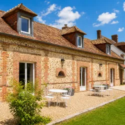 Maison en brique avec toit en tuiles, tables et chaises blanches sur terrasse sous ciel bleu avec nuages.