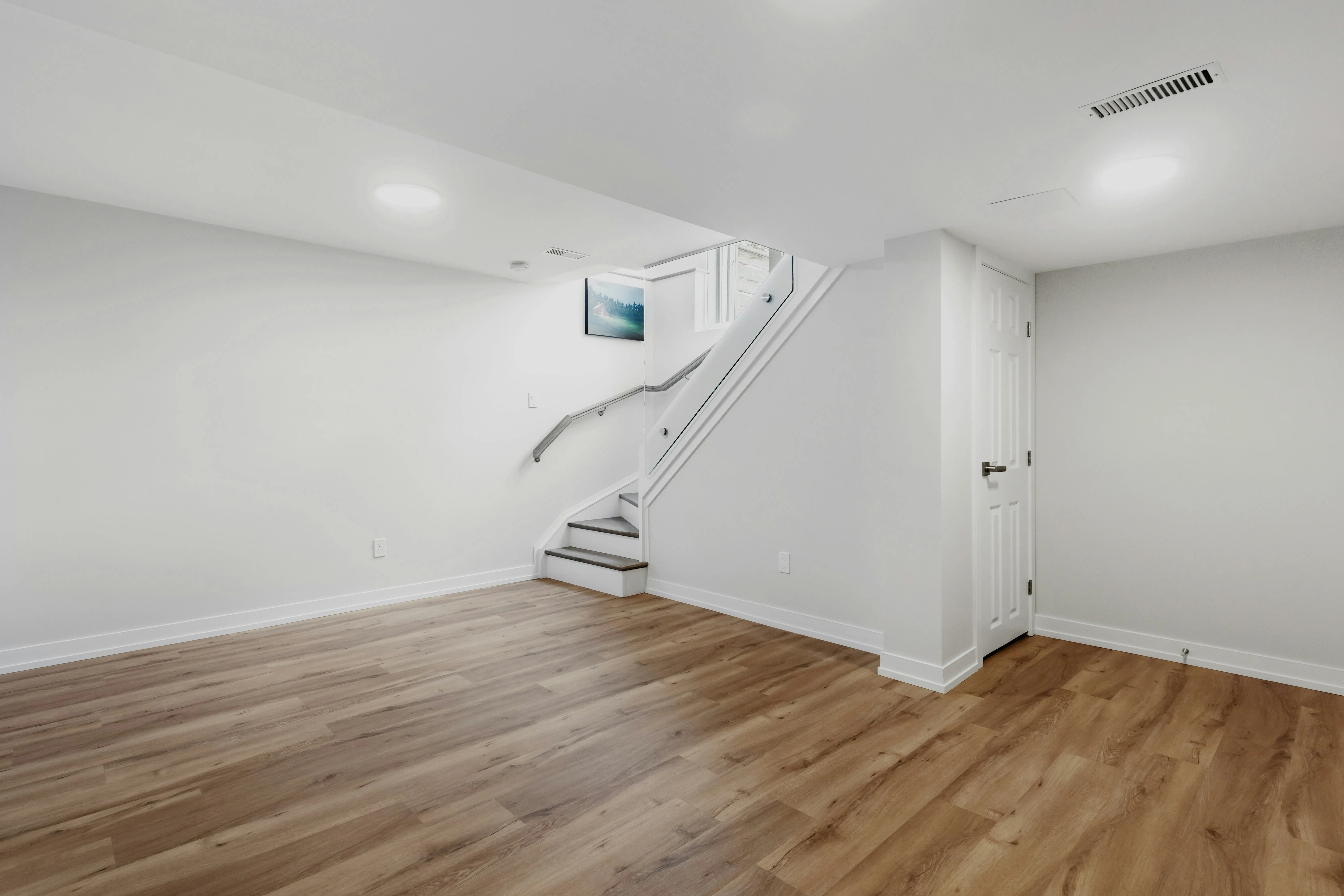Empty basement room with white walls, wooden floor, small staircase with glass railing, and a closed white door.