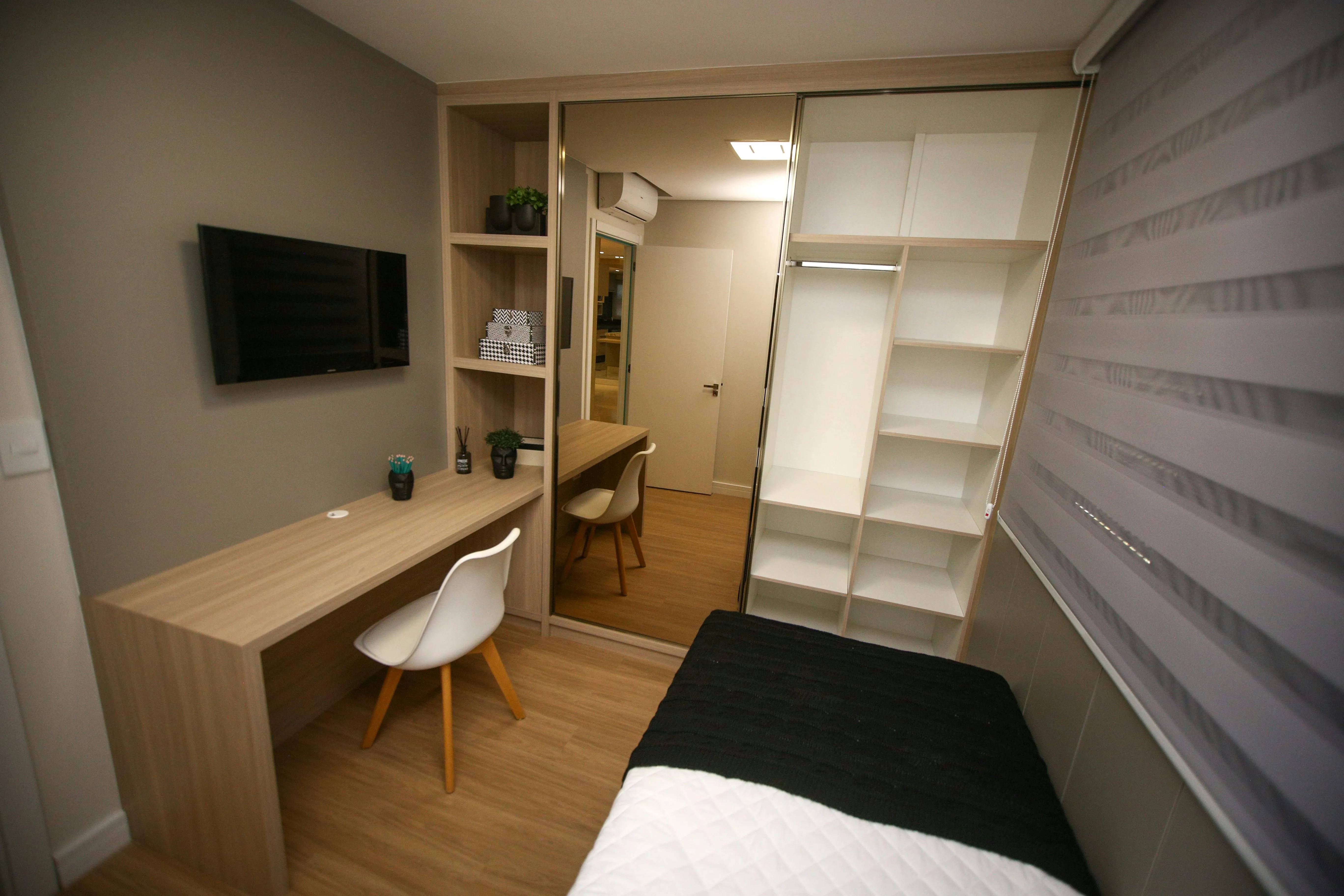 Small modern bedroom featuring a wooden desk with white chair, wall-mounted TV, mirrored closet doors, and a bed with black and white bedding.